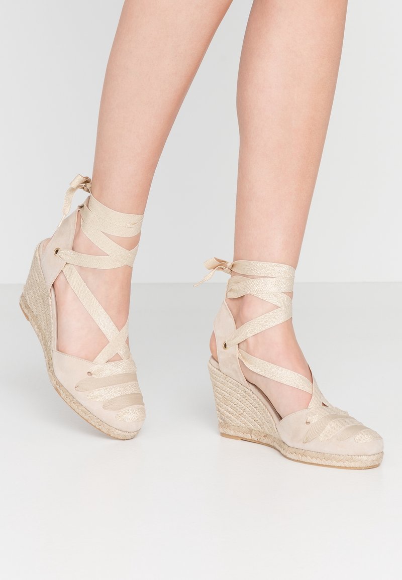 Sandales compensées beiges avec un talon en jute texturé, dotées de brides croisées en ruban et d'une tige en daim avec des accents métalliques.