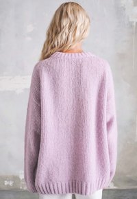 Blondynka w luźnym, jasnofioletowym dzianinowym swetrze stoi tyłem do kamery na tle szarej teksturowanej ściany.