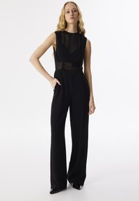 Tuta nera con corpetto trasparente, design senza maniche, scollatura alta e pantaloni a gamba larga. Caratterizzata da linee pulite e uno stile minimalista.