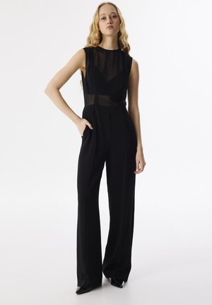 Black jumpsuit with sheer bodice, sleeveless design, high neckline, and wide-leg trousers. Features clean lines and minimalist style.