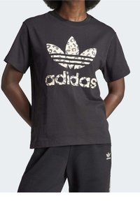 Person som bär en svart Adidas-t-shirt med stort leopardtryck och matchande svarta byxor med litet logo på låret.