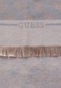 Vékony szövésű pamut takaró világosszürke színben, bonyolult arany mintákkal és fráncokkal az alsó szélen; kék színű "GUESS" logóval.