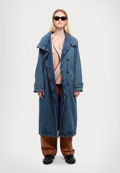 Denim trenchcoat med stor krage, frontknapper og lomme med lapper. Båret over en flerfarget, mønstrete topp og brune vide bukser.