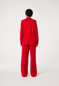 Rode op maat gemaakte pak met een getailleerde blazer en broek met brede pijpen. Gladde stof, minimale details en een gestructureerd rugontwerp.