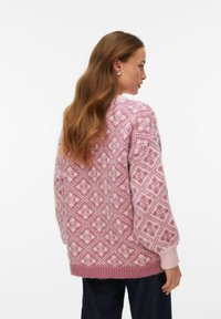 Pull en rose à motif géométrique, avec une coupe ample, une texture épaisse et des poignets côtelés. Porté avec un jean bleu foncé.