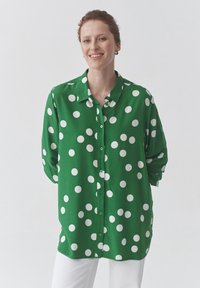 Žalia marškinių tipo palaidinė su baltais taškeliais, ilgomis rankovėmis ir laisvu siluetu. Lygi audinio tekstūra, apykaklė ir priekyje sagos.