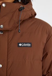 Smeđa izolirana jakna s visokim ovratnikom, koja ima drukere i kapu s uzicom. Uključuje crni Columbia logo na prsima.