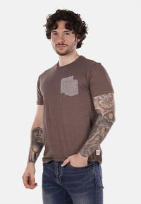 T-shirt in cotone marrone con taschino grigio testurizzato, maniche corte e vestibilità comoda. Indossato con jeans in denim blu.