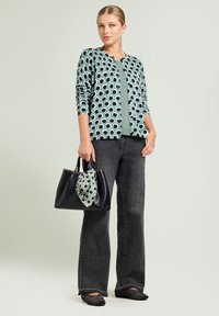 Kobieta w zielonej kardiganie w kropki nosi zielony top, czarne szerokie spodnie, czarne baleriny i trzyma czarną torebkę z pasującym szalem.