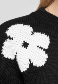 Un pull en tricot noir avec un grand motif de fleur blanche sur l'épaule, en tissu texturé avec un col côtelé.