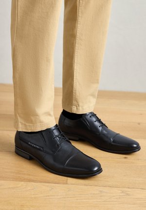 Chaussures de ville en cuir noir avec une finition lisse, présentant un bout arrondi, un design à lacets et un branding subtil sur le côté, associées à un pantalon khaki.