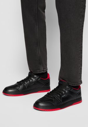 Czarne niskie trampki z czerwonymi podeszwami i wnętrzem, noszone z czarnymi skarpetkami i ciemnoszarymi jeansami na jednolitym jasnoszarym tle.