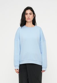 Sweatshirt bleu clair avec un col rond, épaules tombantes et poignets côtelés. Tissu doux, coupe décontractée et texture lisse.