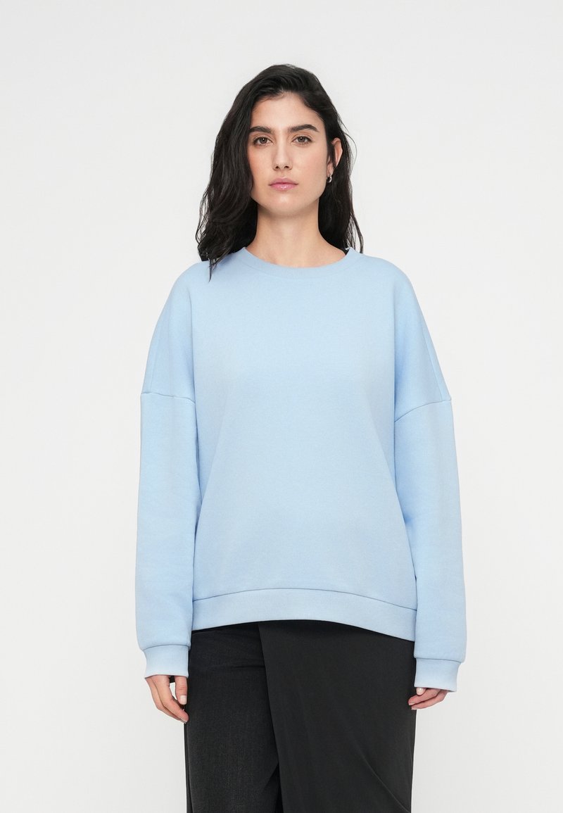 Sweatshirt bleu clair avec un col rond, épaules tombantes et poignets côtelés. Tissu doux, coupe décontractée et texture lisse.