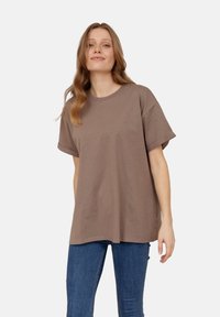 Brązowa bawełniana koszulka z luźnym krojem, krótkimi rękawami i okrągłym dekoltem, wyeksponowana z niebieskimi dżinsami na białym tle.