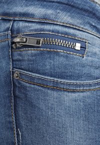 Närbild av en ficka på blå denimjeans med en exponerad silverblixtlås och synliga söm detaljer.