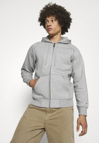 Grauer Reißverschluss-Hoodie mit Kapuze und Kordelzug, Raglanärmeln und Kängurutaschen; aus strukturiertem Baumwollstoff; kombiniert mit beigen Hosen.