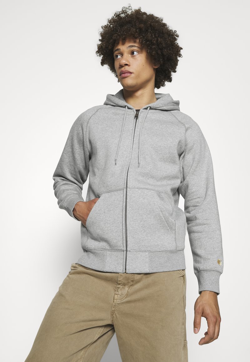 Grauer Reißverschluss-Hoodie mit Kapuze und Kordelzug, Raglanärmeln und Kängurutaschen; aus strukturiertem Baumwollstoff; kombiniert mit beigen Hosen.