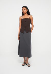 Sieviete stāv, ģērbusies brūnā pogātā bez piedurknēm topā, pelēkā midi svārku un čūsku raksta slidenu apavu fonā uz vienkārša balta fona.