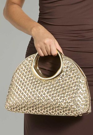 Main d'une femme tenant un grand sac à main tissé doré avec des poignées métalliques circulaires brillantes sur un fond de robe marron plissée.