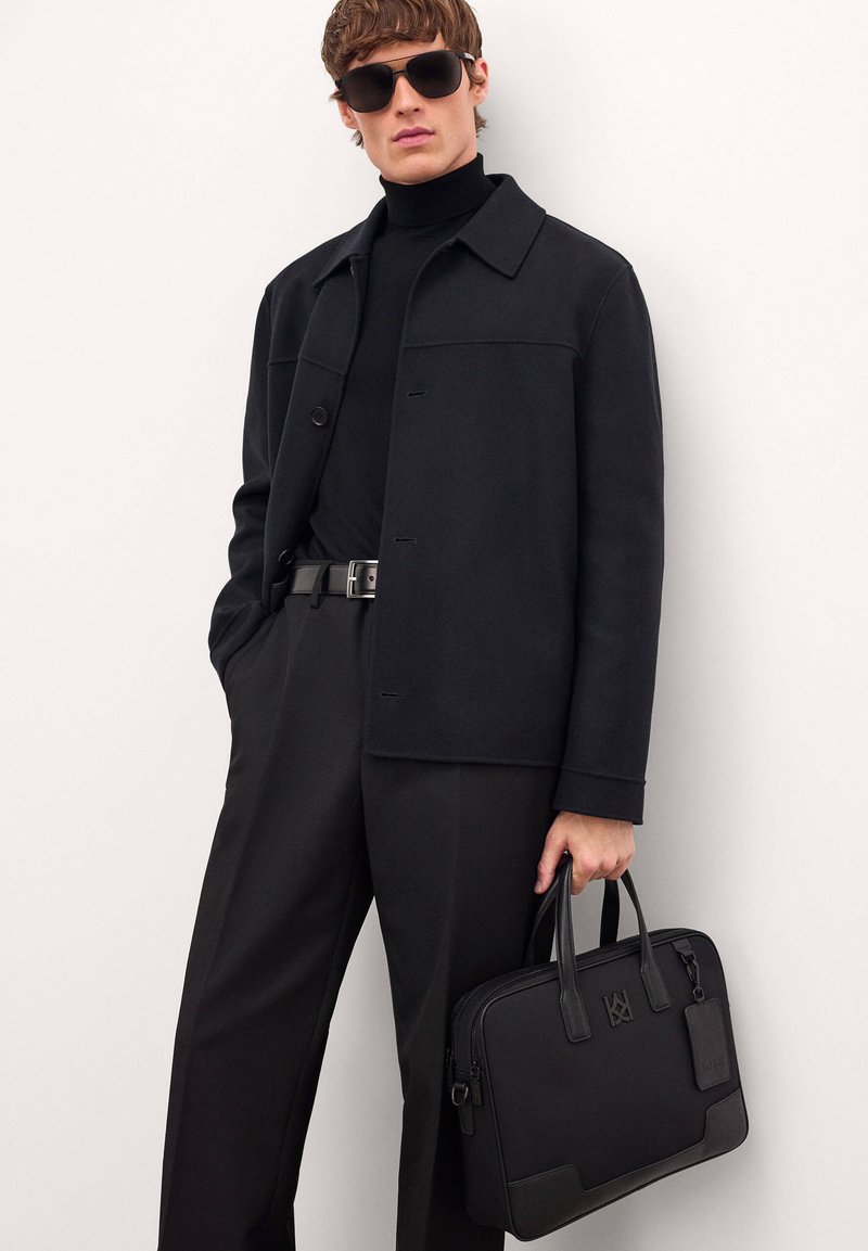 Mustavilla villasekoitteella varustettu takki mustan poolopaidan päällä, yhdistettynä mustiin housuihin. Mallilla on kädessään musta muotilaukku yläkahvoilla.