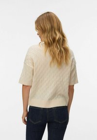 Crèmekleurige gebreide trui met een getextureerd diamantpatroon, korte mouwen en een geribde zoom. Gecombineerd met donkerblauwe jeans.