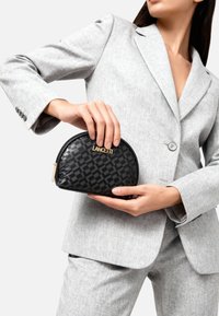 Bustina cosmetica nera con cerniera, decorata con un motivo geometrico testurizzato e un logo dorato, tenuta in mano davanti a un abito grigio sartoriale.