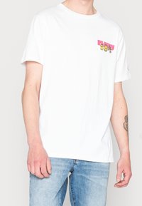 Hvid bomuld t-shirt med korte ærmer og rund hals, prydet med farverig grafisk tekst på den øverste venstre side. Båret med lyseblå jeans.