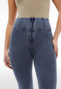 Jeans de mezclilla de cintura alta en azul oscuro con un acabado texturizado, que presentan una cremallera visible y detalles de costura a lo largo de la cintura.