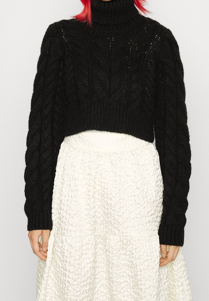 Osoba ubrana w grubą czarną sweter z warkoczowym splotem i wysoko osadzoną białą spódnicę o fakturze, stojąca na tle jednolitym.