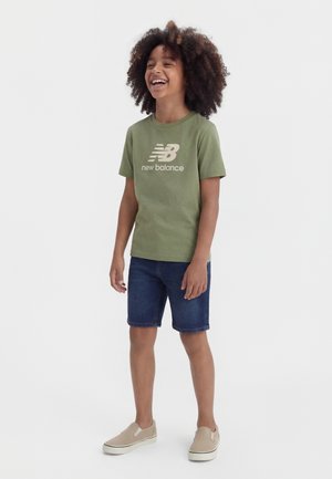 Enfant aux cheveux bouclés portant un t-shirt vert New Balance, un short en denim et des chaussures beige à enfiler, souriant et se tenant devant un fond uni.