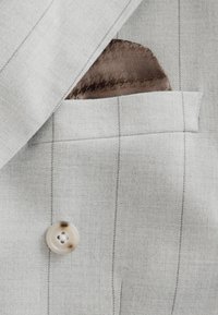 Světlé šedé saka na míru s vertikálními tenkými proužky, s hnědým vzorovaným kapesníčkem a kulatým krémovým knoflíkem.