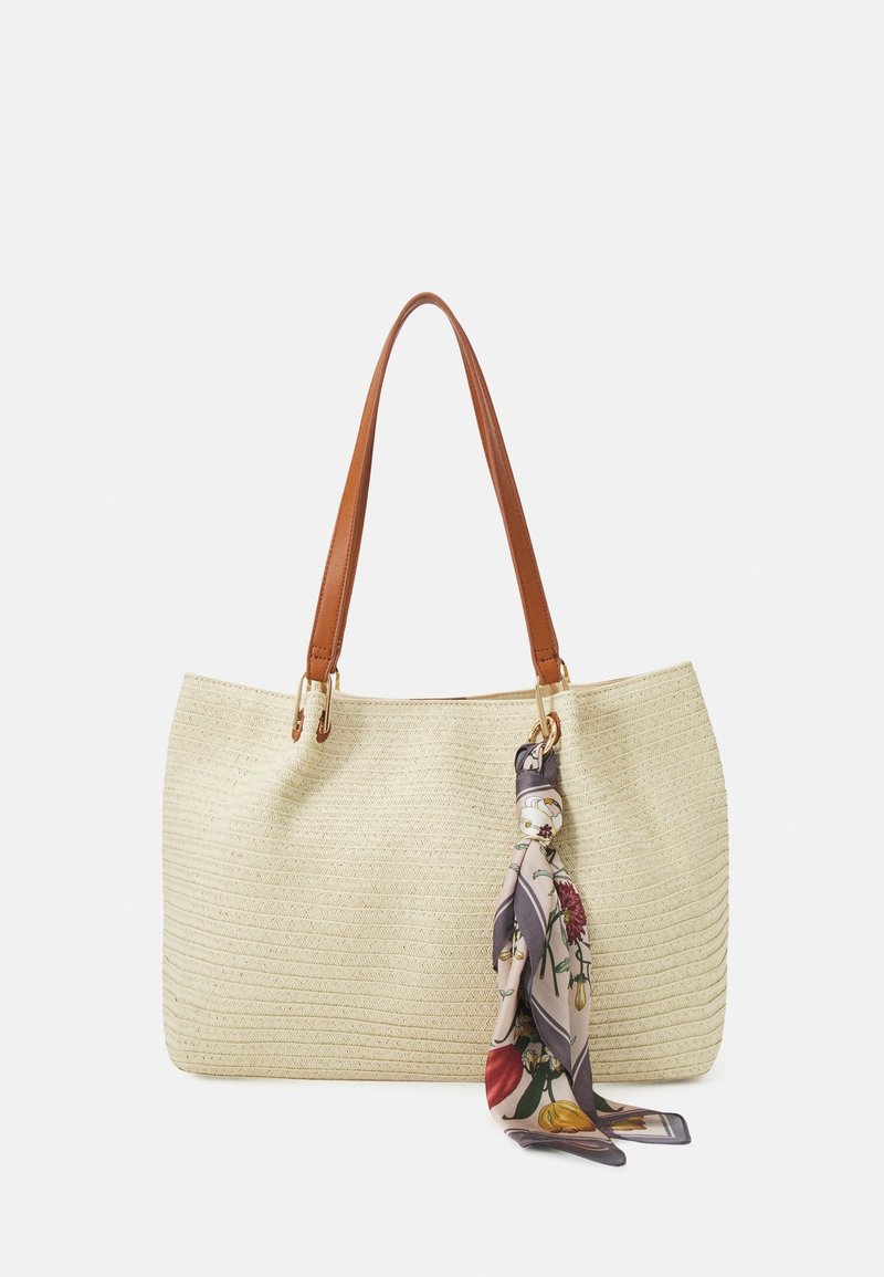 Sac fourre-tout tissé crème avec des poignées en cuir marron et un foulard à motif floral attaché à une poignée, sur fond blanc.