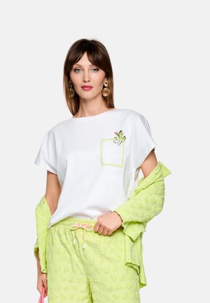 Frau trägt weißes T-Shirt mit grün bestickter Tasche, limettengrüne strukturierte Hose und passende limettengrüne Jacke locker über die Schultern gelegt, hält eine pinke Tasche.