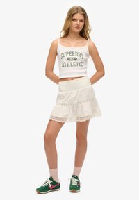 Weißes, kurzes Tanktop mit grünem Slogan, kombiniert mit einem weißen, schichten Rock mit Spitzenrand. Das Model trägt grüne Sneakers mit pinken Akzenten.