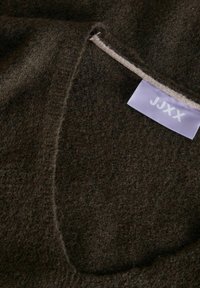Tela marrón oscura con una superficie suave y texturizada. Una etiqueta morada con "JJXX" está cosida en la tela, mostrando un diseño limpio y sencillo.