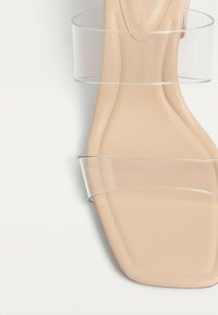 Béžové sandály s plochou podrážkou, transparentním páskem, čtveratou špičkou, hladkým povrchem a minimalistickým designem. Ploché podešve s jemnými detaily.