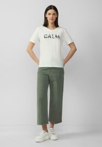 Weißes Baumwoll-T-Shirt mit dem Aufdruck "CALM" in einem gemusterten Design, kombiniert mit khakigrünen, weit geschnittenen Hosen und weißen Sneakers.