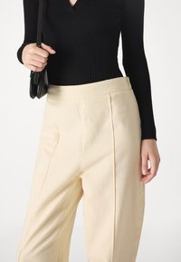 Top lungo a maniche lunghe nero a coste abbinato a pantaloni beige a vita alta con due pieghe frontali. Mani che tengono una piccola borsa nera.