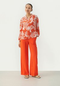 Rød bluse med blomstermønster og lange ærmer, parret med løstsiddende orange bukser med bred ben. Modellen bærer lyse sandaler; glat stof, afslappet pasform.