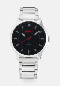 Montre en métal argenté avec un cadran noir, des accents rouges et des marqueurs d'heures. Dispose d'un bracelet en métal poli et d'une résistance à l'eau jusqu'à 30 mètres.