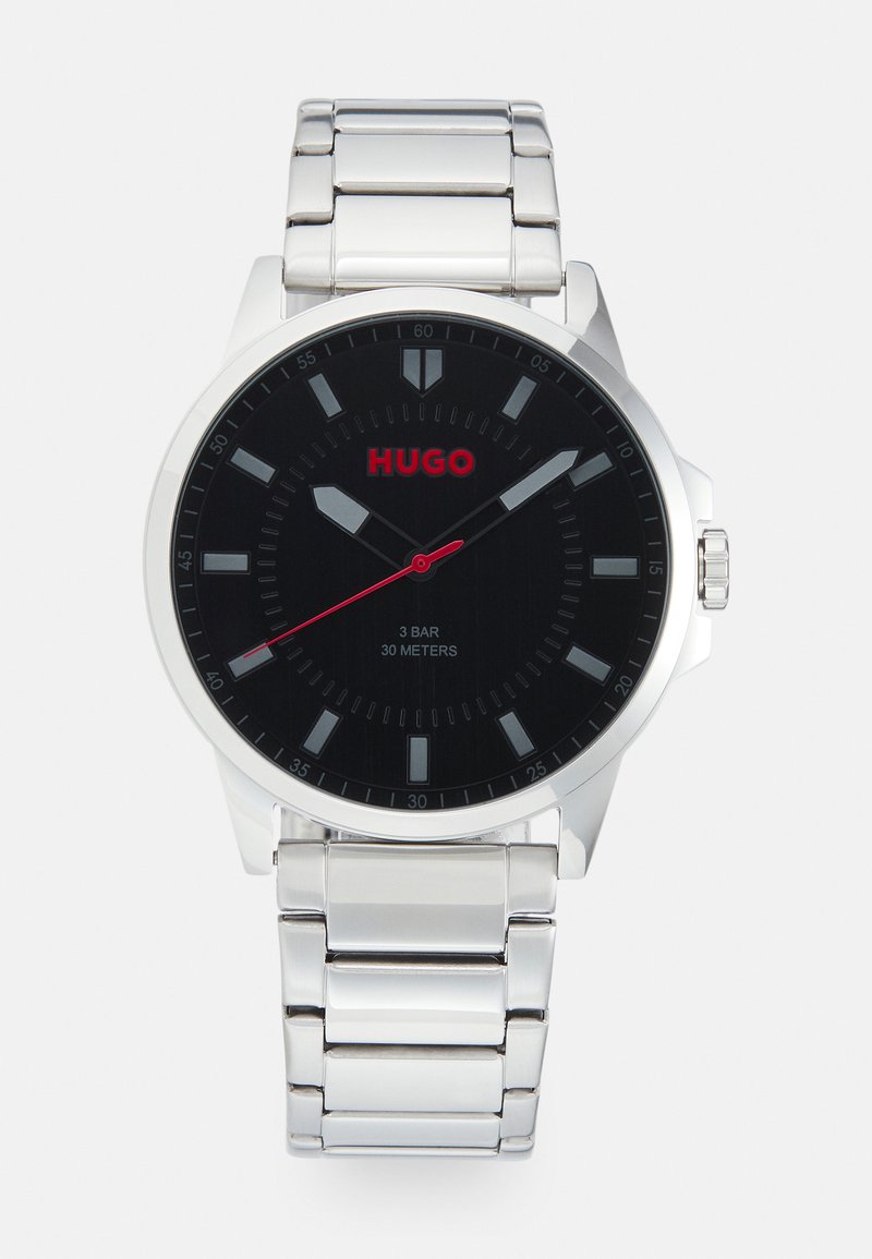 Montre en métal argenté avec un cadran noir, des accents rouges et des marqueurs d'heures. Dispose d'un bracelet en métal poli et d'une résistance à l'eau jusqu'à 30 mètres.