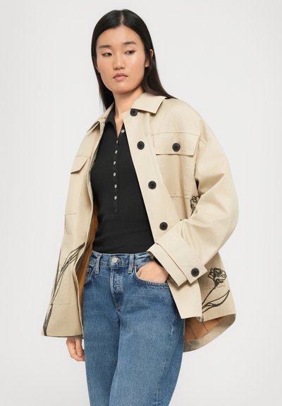 Beige jacket with black buttons and floral embroidery, paired with a black top and blue denim jeans. The jacket features a relaxed fit and front pockets.