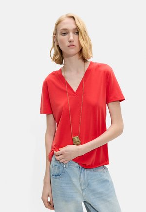 Młoda kobieta o krótkich blond włosach, ubrana w czerwoną koszulkę z dekoltem w serek, jasnoniebieskie dżinsy i długi złoty naszyjnik z zawieszką.