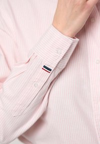 Približan posnetek roke, ki prilagaja manšeto svetlo roza črtaste srajce z belimi gumbi in majhno rdečo, belo in modro zastavo.