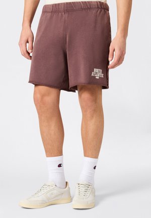 Hombre vistiendo pantalones cortos marrones con cintura elástica y texto "North Atlantic", calcetines blancos con logo de Champion, y zapatillas casuales color blanco roto, de pie sobre un suelo gris.