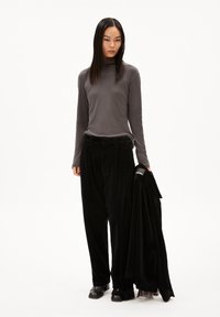 Langärmliges, graues Turtleneck-Oberteil kombiniert mit lockeren, schwarzen Cordhosen. Das Model hält eine schwarze Jacke und steht vor einem weißen Hintergrund.