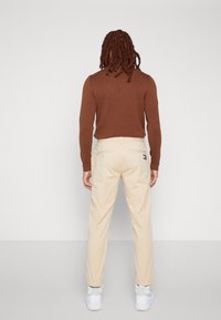 Beige trousers with a straight-leg design, paired with a brown long-sleeve top. The pants feature a back pocket and subtle branding detail.
