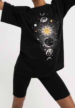 Schwarzes oversized T-Shirt mit einem weißen und goldfarbenen himmlischen Motiv mit Sonne und Monden, kombiniert mit schwarzen Radlerhosen.