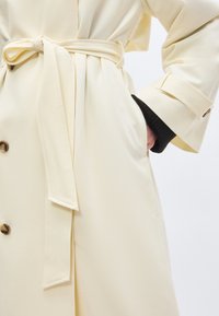 Μπεζ παλτό τύπου τραντσό με ζώνη δεμένη στη μέση, λεπτομέρειες με κουμπιά, ιμάντες στις μανσέτες και ένα χέρι που έχει τοποθετηθεί στην πλευρική τσέπη πάνω από μαύρο μανίκι.